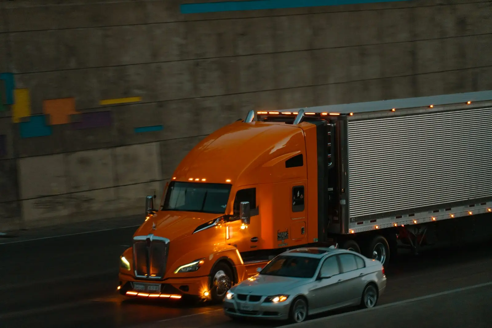 An orange semi-truck and a silver sedan driving on a highway at night