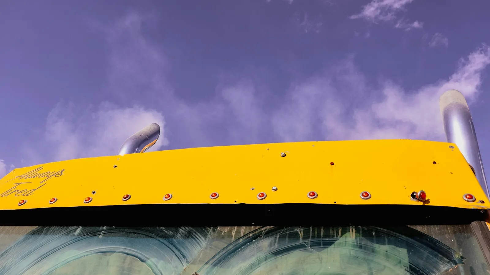 Primer plano de la parte superior de un camión amarillo industrial visto desde ángulo bajo, con luces metálicas y tubos de escape contrastando contra un cielo a